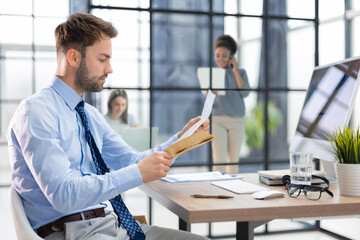 Handsome entrepreneur reading a letter from envelope in a desktop at office with collegues on the background.