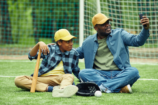 African father in cap making selfie on his mobile phone together with his son while they sitting on grass on field after baseball