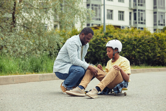 African dad putting protection on knees to his son before skateboarding outdoors - Powered by Adobe