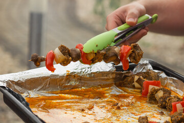Man's hand holding with green forceps a raw meat skewers in spicy marinade. Ready to cook on grill.