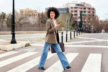 smiling young business woman walking on a crosswalk using her smart phone, concept of communication and urban lifestyle - obrazy, fototapety, plakaty