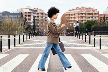 smiling latin business woman walking on a crosswalk using her smart phone, concept of communication and urban lifestyle