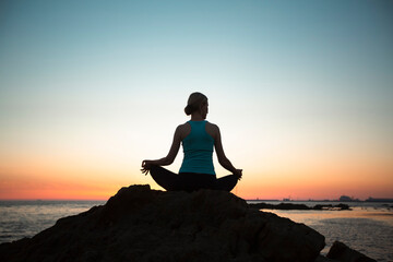 Woman, practicing yoga in lotus pose on the coast during sunset.