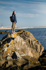Fototapeta na wymiar A woman tourist with a backpack stands on the sea coastal rocks.