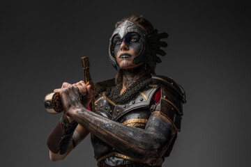 Attractive valkyrie from past with make up holding sword on her shoulder against grey background.
