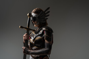 Shot of dramatic female warrior with sword dressed in steel armor posing agianst grey background. - obrazy, fototapety, plakaty