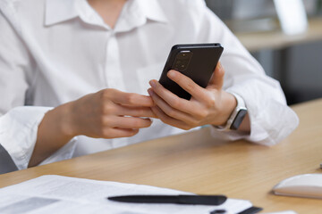 Close-up photo. The hands of a young woman in a white shirt are holding a mobile phone, typing a message at the table.