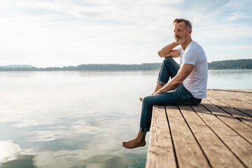 Thoughtful mature man sitting at the edge of pier by lake