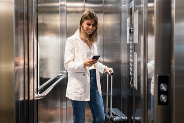 Smiling businesswoman using smart phone standing with suitcase in elevator