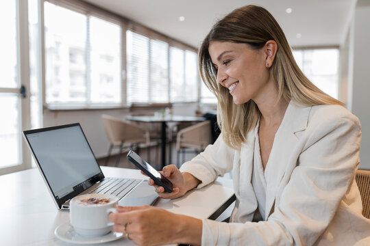 Smiling businesswoman holding mobile phone and coffee cup sitting at cafe