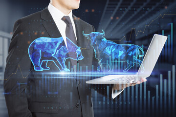  bulls and bears struggle. Equity market illustration. Close up of businessman hand holding laptop with creative hologram and graph on blurry office interior background. Double exposure.