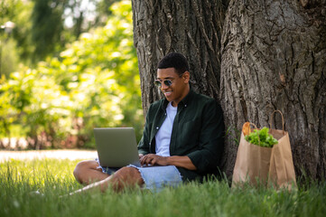 Guy typing on laptop sitting leaning on tree