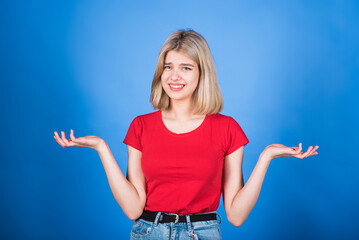 Young and attractive caucasian blonde girl in casual clothes is confused and smirking, shrugging her shoulders isolated on a blue studio background.