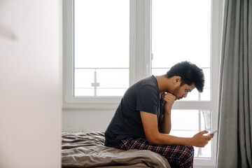 Young handsome indian man sitting on bed with phone
