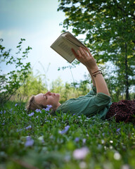 Young woman lying on the grass in the meadow full of violets and reading poetry