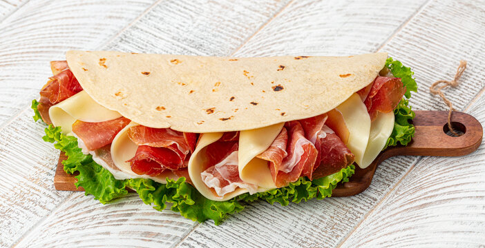Piadina romagnola on a wooden cutting board, traditional italian flatbread with prosciutto crudo di Parma - italian dry cured ham, mozzarella cheese, lettuce.