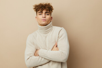  a handsome man with curly hair, standing on a beige background in a light turtleneck and looking at the camera with his arms crossed on his chest. Photo with space for inserting an advertising layout