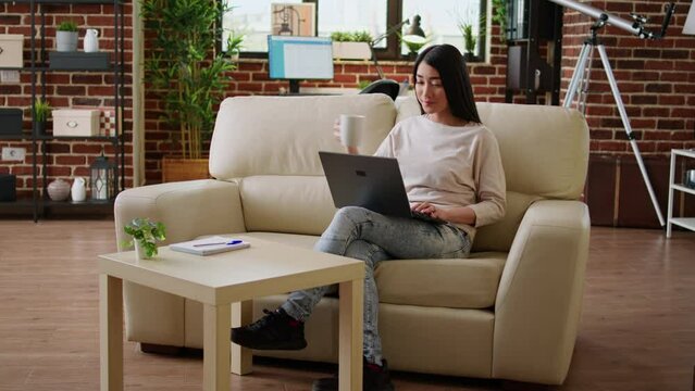 Focused young adult asian student attending online class while typing important lessons on modern portable computer at home. Beautiful woman sitting on sofa while working remotely on laptop.