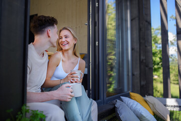 Young couple sitting, drinking coffee and cuddling in hammock terrace in their new home in tiny...