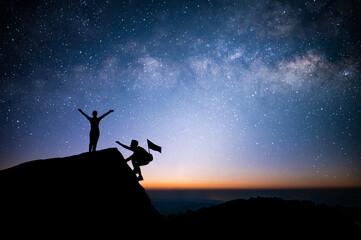 Two young tourist with backpack is climbing a cliff with beautiful night sky, Milky Way and star over the sky. He had an effort to climb all the way to the top of the mountain and he had to succeed.
