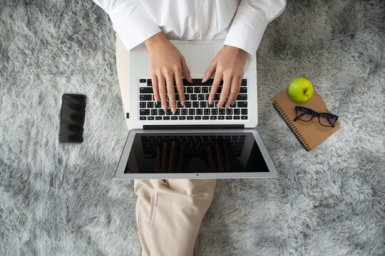 Flat lay, top view of young woman sitting on carpet and using laptop for online learning with notebook, smartphone, eyeglasses and green apple placed on ground in living room at home.