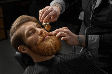 Barbershop Shaving. Male barber shaves beard. Close-up of young bearded man getting beard haircut by hairdresser or barber at barbershop. - obrazy, fototapety, plakaty