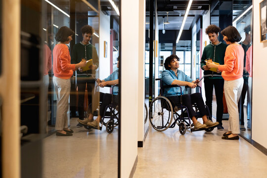 Multiracial colleagues discussing with young businessman sitting in wheelchair at office corridor