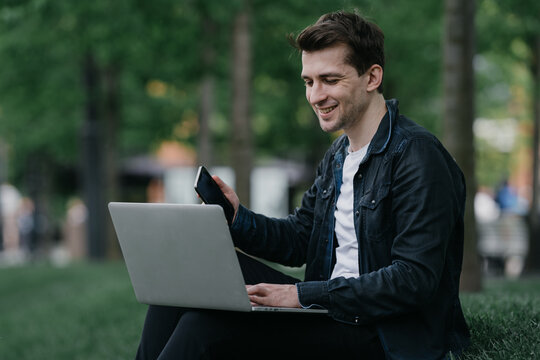 Cheerful Young Man Working At Park Sitting On Lawn, Making Video Call With Friend. Student Making Distant Call, Talking To Parents Living Far. Young Entrepreneur Happy To See Company's Profit Growing.