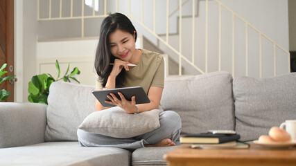 Smiling young asian woman in casual clothes using digital tablet on couch in cozy living room