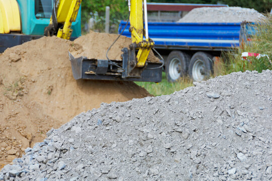 Bodenaustausch - Bagger mit vollem Baggerl&ouml;ffel und Schotterhaufen im Vordergrund