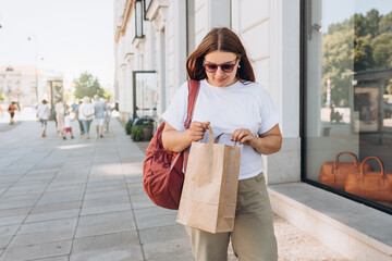Beautiful fashionable young woman in sunglasses with shopping paper bag standing on city street, black friday. Urban lifestyle concept. Delivery carrying.