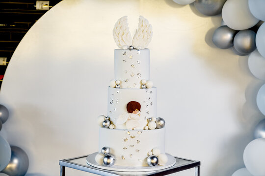 White christening cake with angel wings on top in banquet hall with the balloons behind