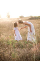 Fototapeta na wymiar A cute little girl with long blond curly hair and her mother in a white summer dress and a straw boater hat in a field in the countryside in summer at sunset. Nature and Ecolife