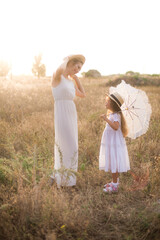 Naklejka na ściany i meble A cute little girl with long blond curly hair and her mother in a white summer dress and a straw boater hat in a field in the countryside in summer at sunset. Nature and Ecolife