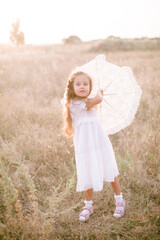 A cute little girl with long blond curly hair and in a white summer dress and a straw boater hat in a field in the countryside in summer at sunset. Nature and Ecolife