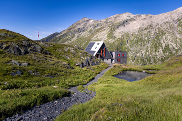 Fototapeta na wymiar View of the Scaletta hut near the Greina Pass in Blenio, Switzerland. In the foreground is the path leading to the hut. In the background are the high mountains overlooking the scene.
