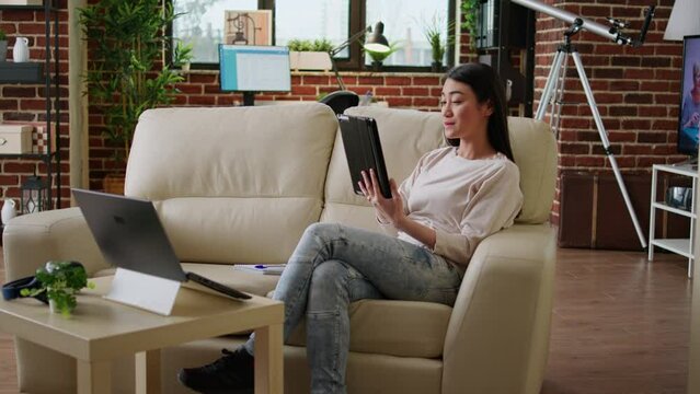 Happy asian woman having modern tablet browsing internet while sitting on couch at home. Cheerful smiling heartily person with touchscreen device surfing webpages while doing remote work.