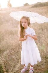 A cute little girl with long blond curly hair and in a white summer dress and a straw boater hat in a field in the countryside in summer at sunset. Nature and Ecolife