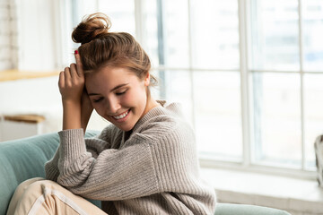 Portrait of sweet smiling young woman in homewear sitting on nice sofa at home. Morning and lifestyle concept. Copy space in right side. Window on blurred background