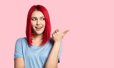 Portrait of charming smiling lady pointing away with finger. Red-haired attractive young female in blue shirt. Positivity and happiness concept. Isolated on pink background