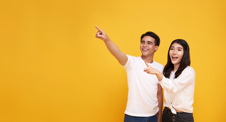 portrait of happy excited Asian couple tourists pointing hands to empty space on isolated light yellow background.