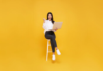 Young woman asian happy smiling. While her using laptop sitting on white chair and looking isolate on yellow background.
