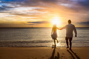 A romantic holiday couple holding hands enjoys a beach during golden sunset time and runs in the sea