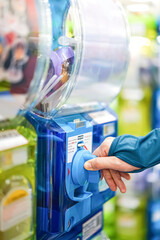 A human hand in a turquoise sweater is twisting a blue gashapon machine to release a figure. In Japan, it is considered as one of the most well liked risks.