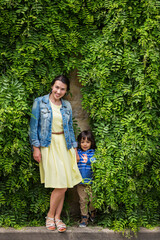 Fototapeta premium Woman and little handsome baby boy staying surrounded by lush green plants, touching leaves. Young woman and kid near leaves wall
