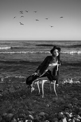 girl in stylish formal suit, shirt and hat sitting on white chair in water of ocean.  Romantic vertical black and white composition. loneliness. love and hope metaphor waiting by the sea. 
