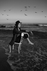  Romantic melancholic black and white horizontal composition loneliness by the sea. girl in a formal elegant stylish suit, shirt and hat sitting on chair in waves of the sea. artistic dreamer portrait
