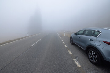 Fototapeta na wymiar A gray SUV car is parked on the side of a straight empty road invaded by thick fog. In the background, the silhouettes of some tall trees, shrouded in fog, can be guessed.