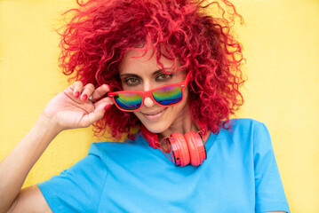 close-up of a smiling young Latin woman with red afro hair wearing a blue t-shirt, looking behind sunglasses at the camera on yellow background, red headphones around her neck