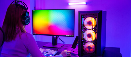 Asian professional gamer playing online video game on desktop computer PC have colorful neon LED lights dark room, young woman in gaming headphones using computer for playing game at home, Back view